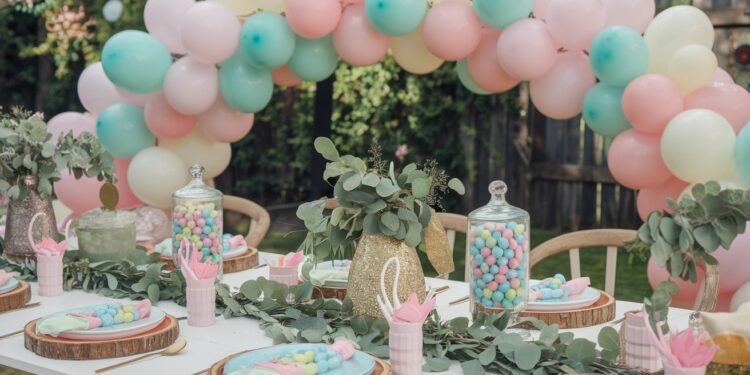 A festive outdoor party table setup with pastel pink, blue, and white balloons forming an arch. The table is adorned with glass jars filled with colorful candies, green foliage, and pink napkins under clear skies.