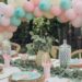 A festive outdoor party table setup with pastel pink, blue, and white balloons forming an arch. The table is adorned with glass jars filled with colorful candies, green foliage, and pink napkins under clear skies.