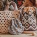 A display of three handcrafted bags in various crochet and woven designs on a wooden surface. Yarn balls, crochet needles, and dried flowers are scattered around, with patterned fabric in the background.