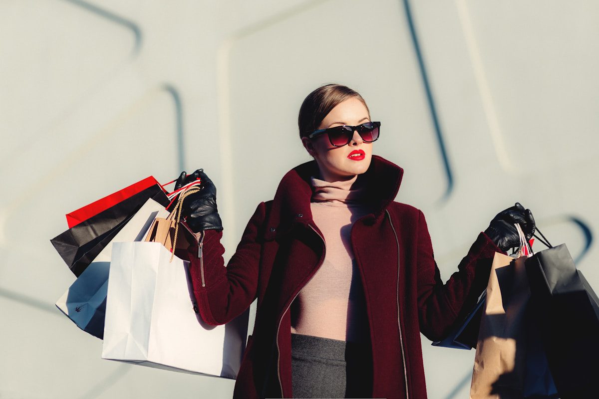 Chic woman in sunglasses, burgundy coat, and black gloves carries Amazon Best Finds bags outdoors against a sleek light background.